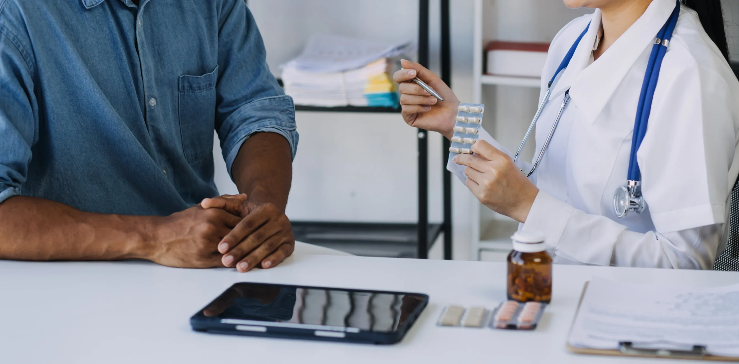 Doctor in a white coat shows blister packs to a patient during a consultation; a tablet and a pill bottle are on the table.