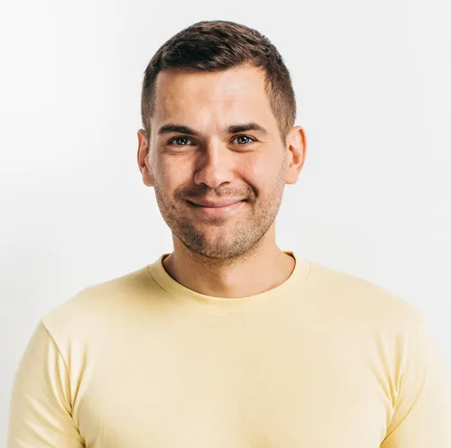 A headshot of Oliver R. with short brown hair and light stubble, wearing a pale yellow t-shirt, smiling subtly against a plain light grey background.