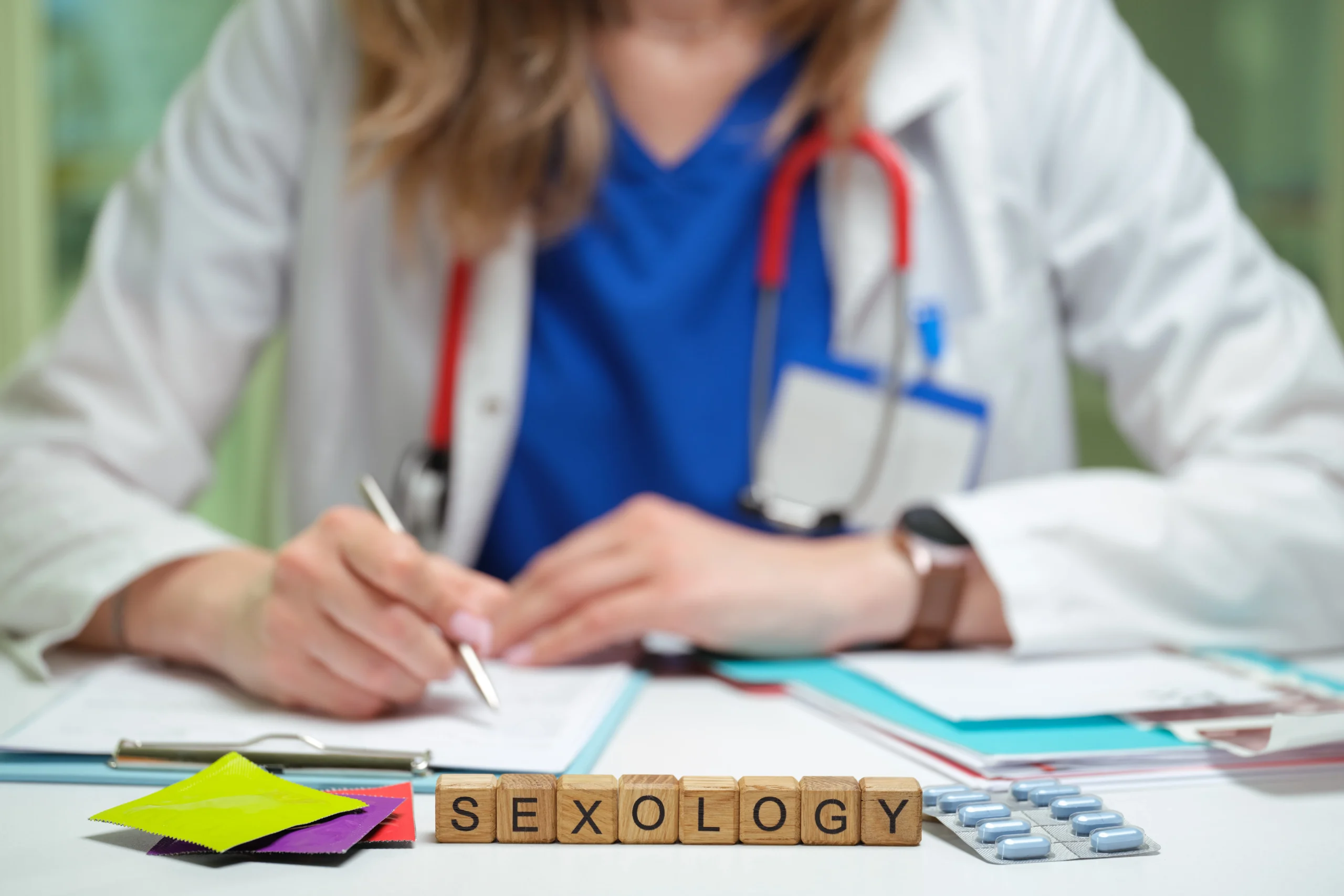 Medical professional writing at a desk, with wooden blocks spelling SEXOLOGY in the foreground.