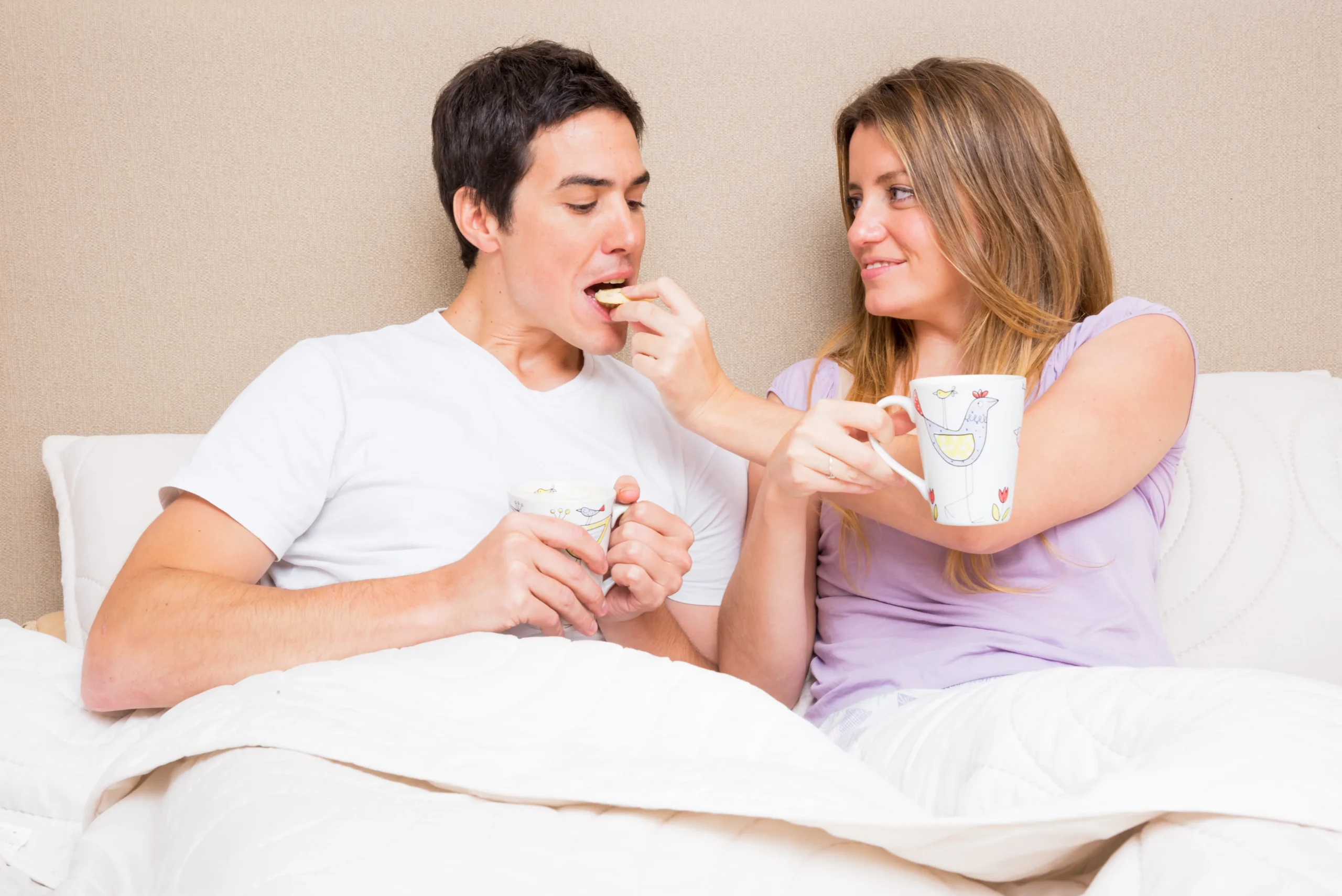 A woman playfully feeds a snack to a man while lounging in bed, reflecting health management and daily PrEP treatment routines during personal moments.