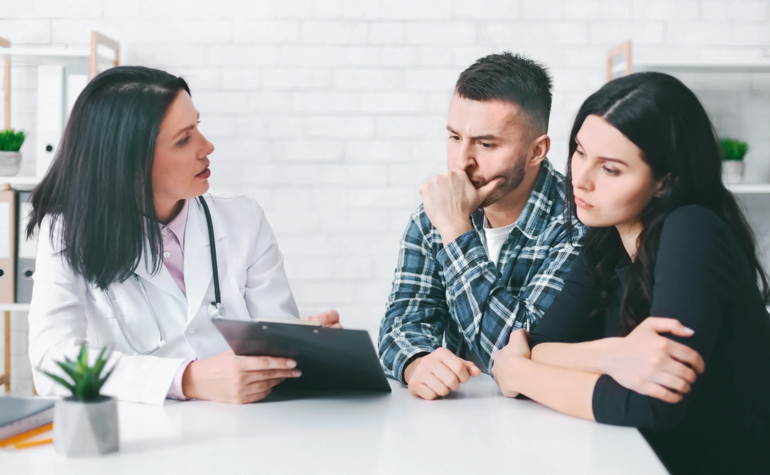 A female doctor in a white lab coat with a stethoscope holding a clipboard and speaking with a concerned young couple sitting across from her at a desk.