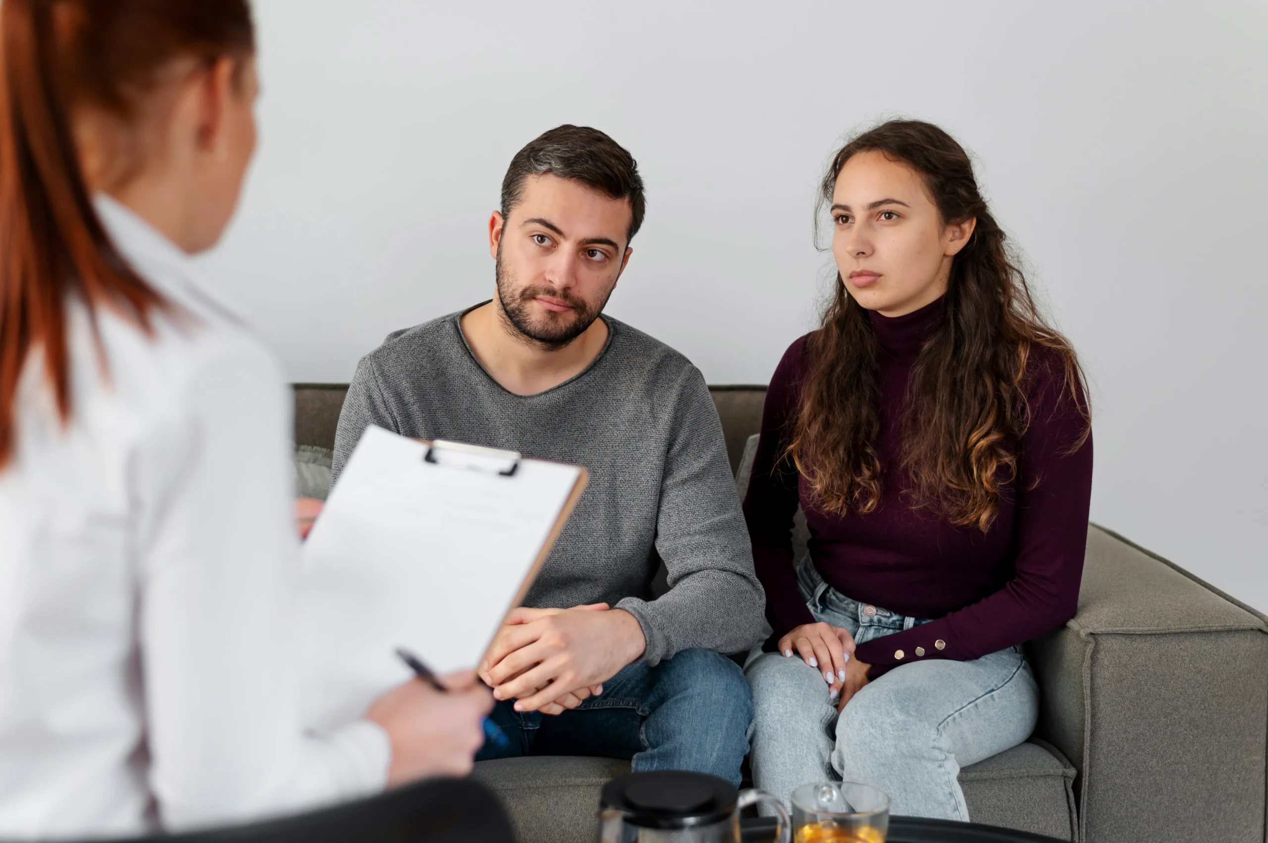 A young couple sitting on a couch listening to a doctor holding a clipboard during a consultation for HIV PrEP.