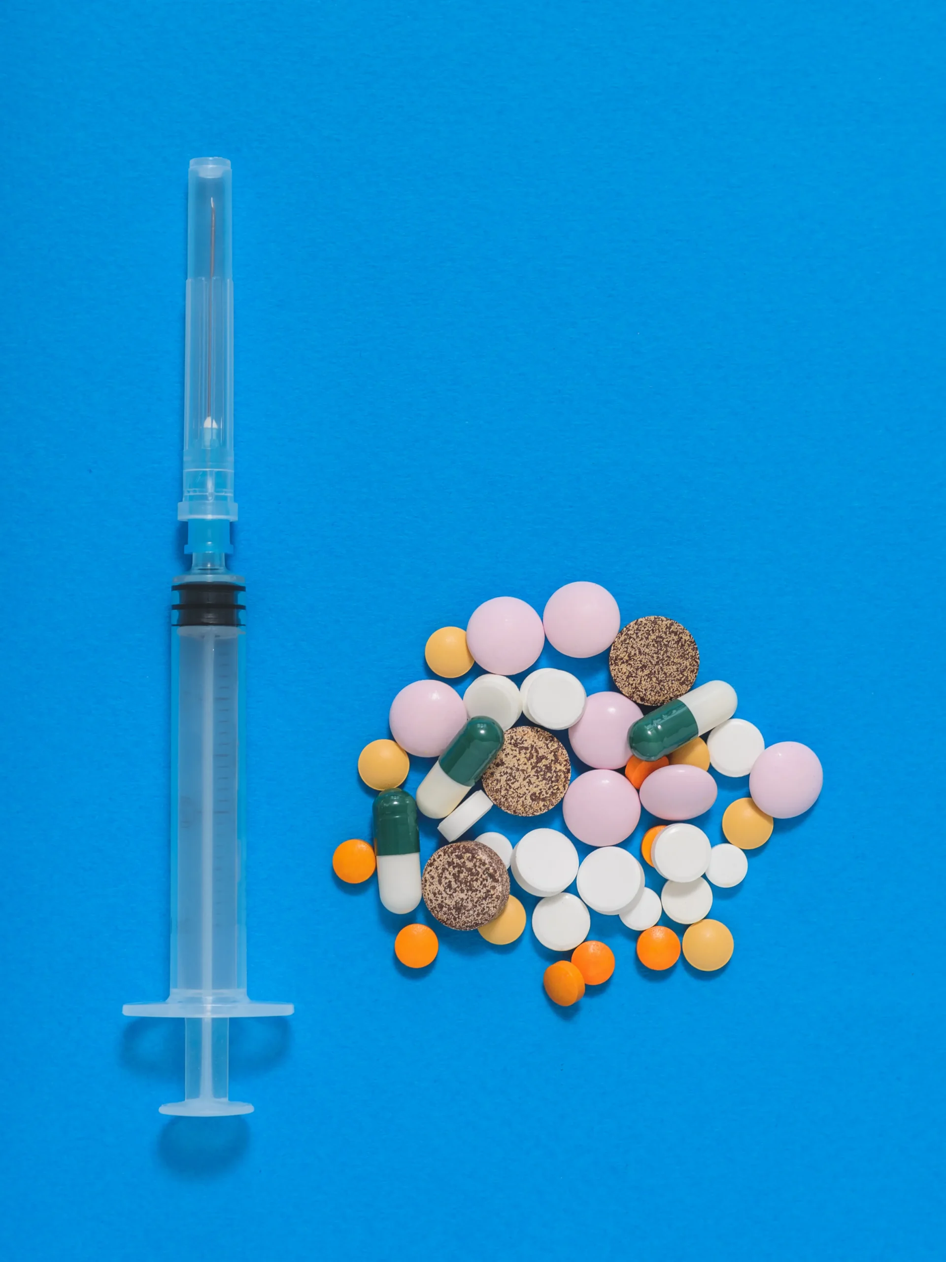 A medical syringe placed next to a diverse pile of colorful prescription medications, illustrating the choice between injectable PrEP (Apretude) and daily oral PrEP pills for HIV prevention.